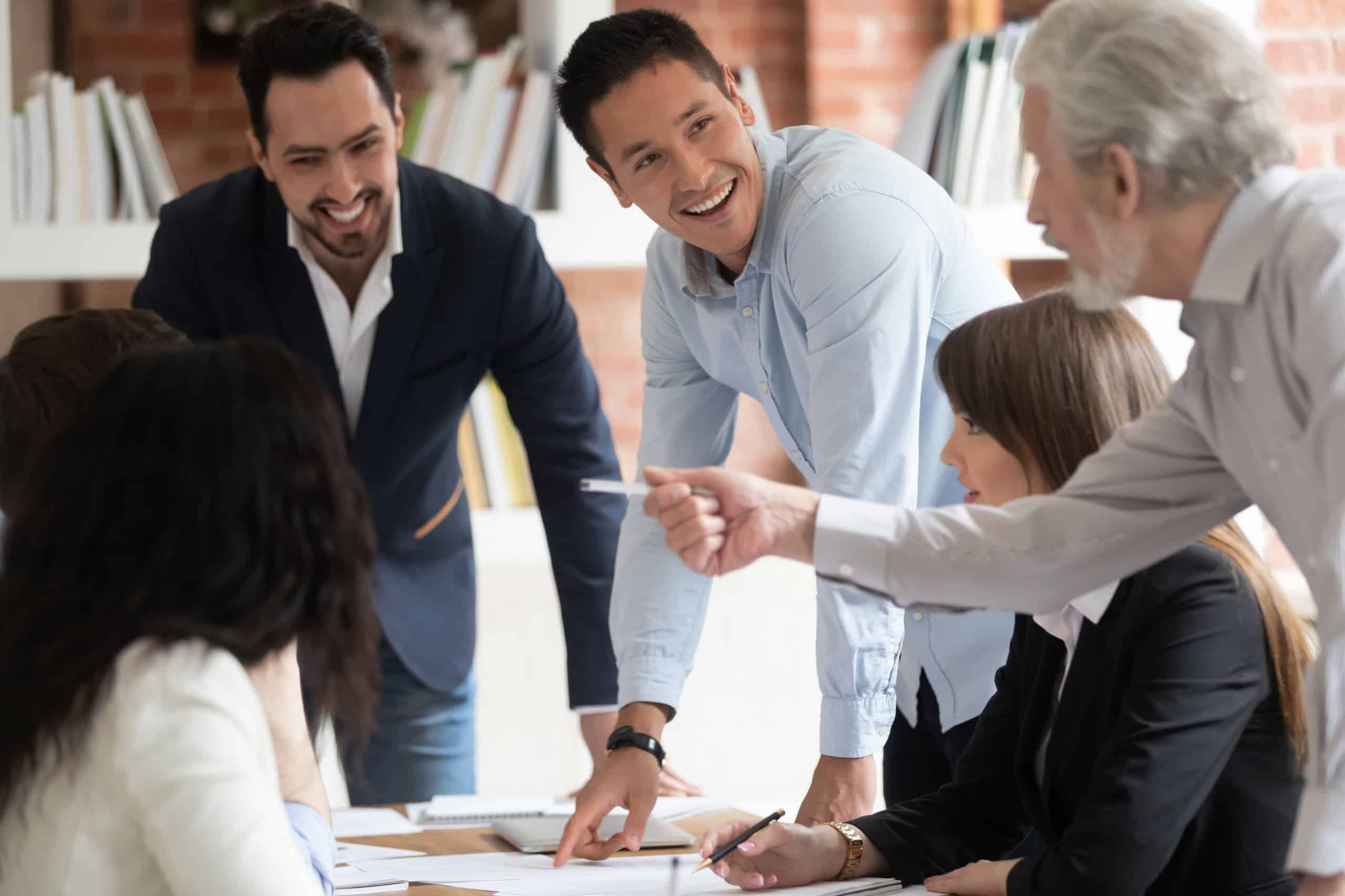 A group of business people gathered sitting and standing around a desk, listening to their colleagues' discussion