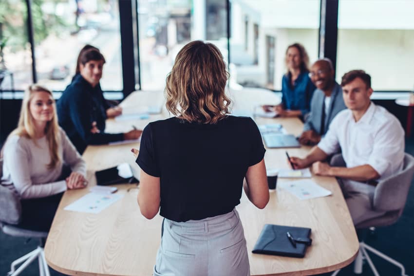 Blonde woman is standing in front of five people who are sitting at a meeting room table