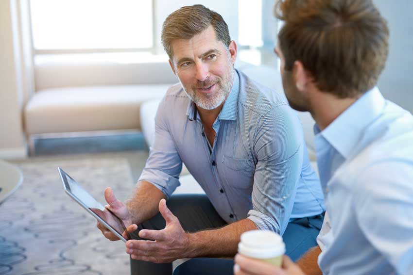 An older man holding a tablet is speaking with a younger colleague with a coffee cup. They sit next to each other on a couch.