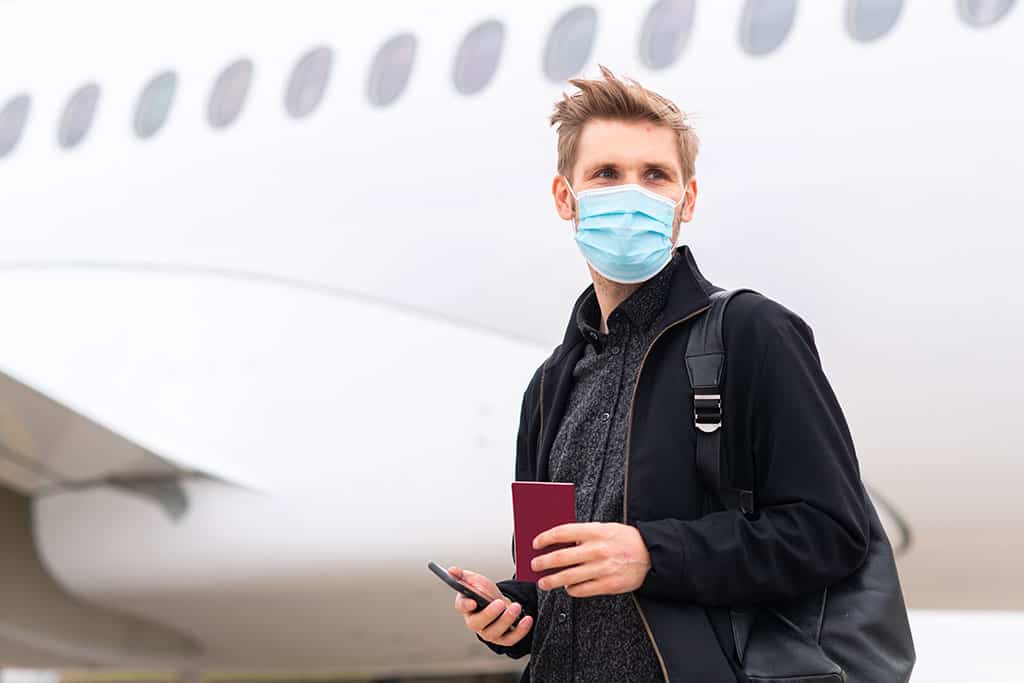 uomo con mascherina e passaporto davanti ad aeroplano