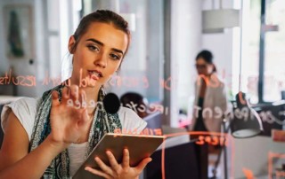 ragazza che scrive con un pennarello su una lavagna trasparente con dietro una collega che annuisce