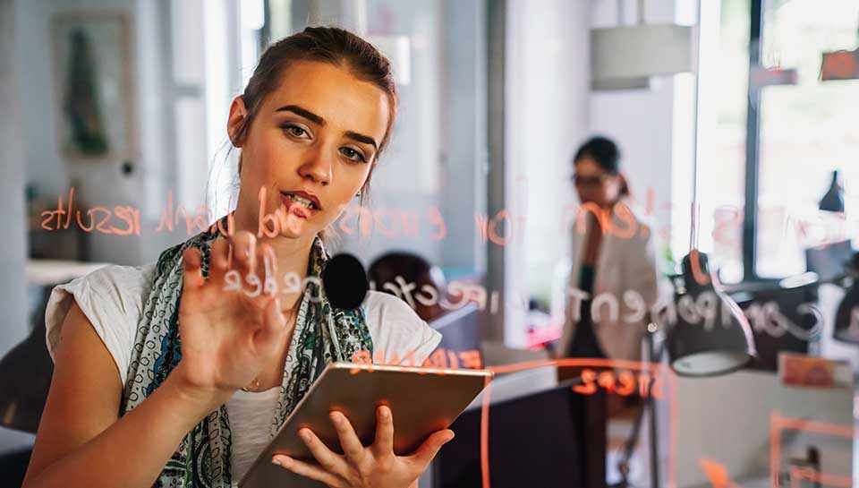 ragazza che scrive con un pennarello su una lavagna trasparente con dietro una collega che annuisce