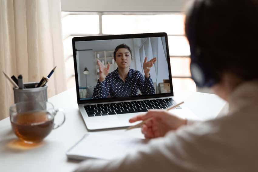 A person in front of a laptop looking at their colleague speaking in a video call and taking notes
