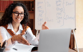 docente che fa lezione online con dietro una lavagna con i tempi verbali inglesi