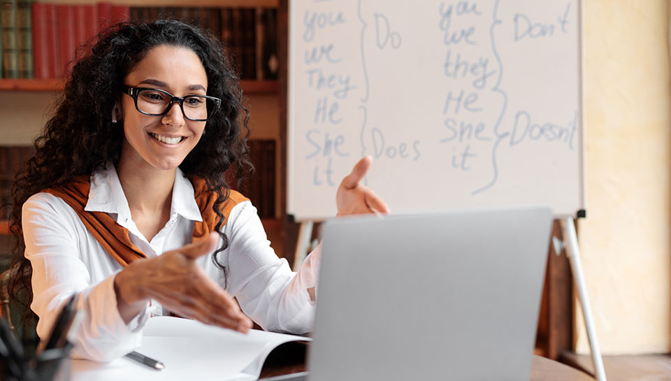 docente che fa lezione online con dietro una lavagna con i tempi verbali inglesi